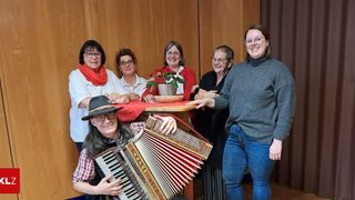 Eine Gruppe lächelnder Frauen und ein Mann mit einem Akkordeon stehen hinter einem Tisch mit einem Topf auf ihm.