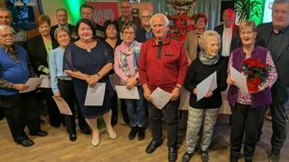 Eine Gruppe von Menschen steht zusammen, hält Urkunden und Papiere und lächelt für ein Foto in einem Raum mit Holzboden und einem geschmückten Weihnachtsbaum im Hintergrund.