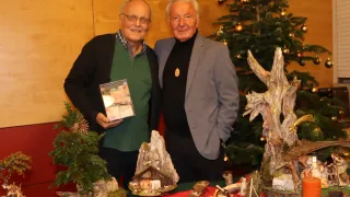Zwei ältere Männer stehen nebeneinander und lächeln. Ein Mann hält eine CD-Hülle mit Geld darin. Sie stehen vor einer Weihnachtsdekoration mit einem kleinen Haus und Baum.