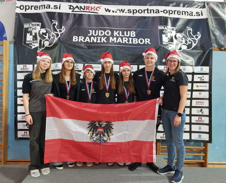 Eine Gruppe von Menschen mit Weihnachtsmützen posiert für ein Foto, während sie eine Flagge mit einem goldenen Adleremblem halten. Hinter ihnen zeigt ein Banner die Worte Judo Klub Branik Maribor.