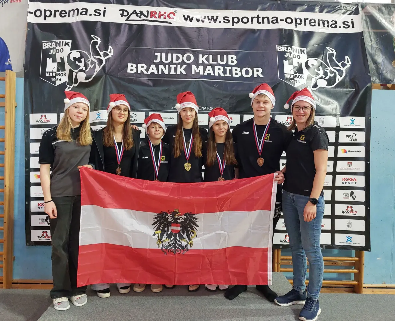 Eine Gruppe von Menschen mit Weihnachtsmützen posiert für ein Foto, während sie eine Flagge mit einem goldenen Adleremblem halten. Hinter ihnen zeigt ein Banner die Worte Judo Klub Branik Maribor.