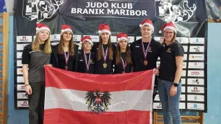 Eine Gruppe von Menschen mit Weihnachtsmützen posiert für ein Foto, während sie eine Flagge mit einem goldenen Adleremblem halten. Hinter ihnen zeigt ein Banner die Worte Judo Klub Branik Maribor.