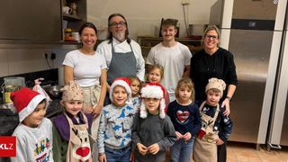 Eine Gruppe von Erwachsenen und Kindern posiert für ein Foto in einer Küche. Sie tragen alle Weihnachtsmützen und Schürzen. Die Küche hat Regale mit Gegenständen und einen Kühlschrank.