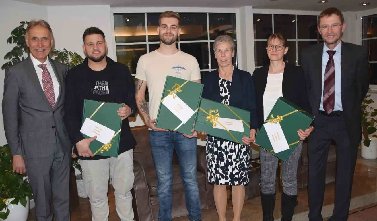 Fünf Personen stehen und lächeln, während sie grüne Geschenkboxen mit weißen Bändern halten. Hinter ihnen steht ein Sofa. Ein weißer Vorhang hängt an der Wand. Einige Lichter reflektieren auf den Glasfenstern.