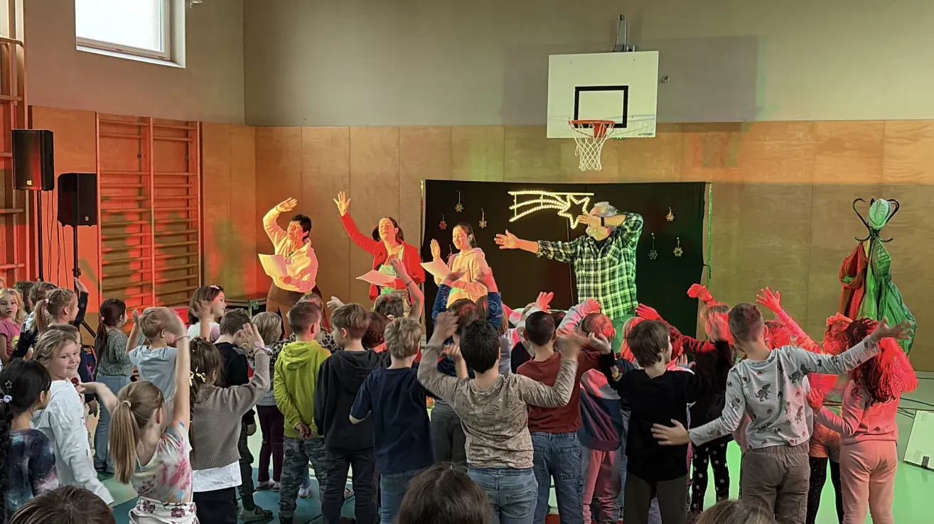 Eine Gruppe von Kindern und Erwachsenen führt ein Theaterstück in einer Turnhalle auf. Die Kinder stehen vorne, während die Erwachsenen auf einer Bühne stehen, mit einem Basketballkorb im Hintergrund.
