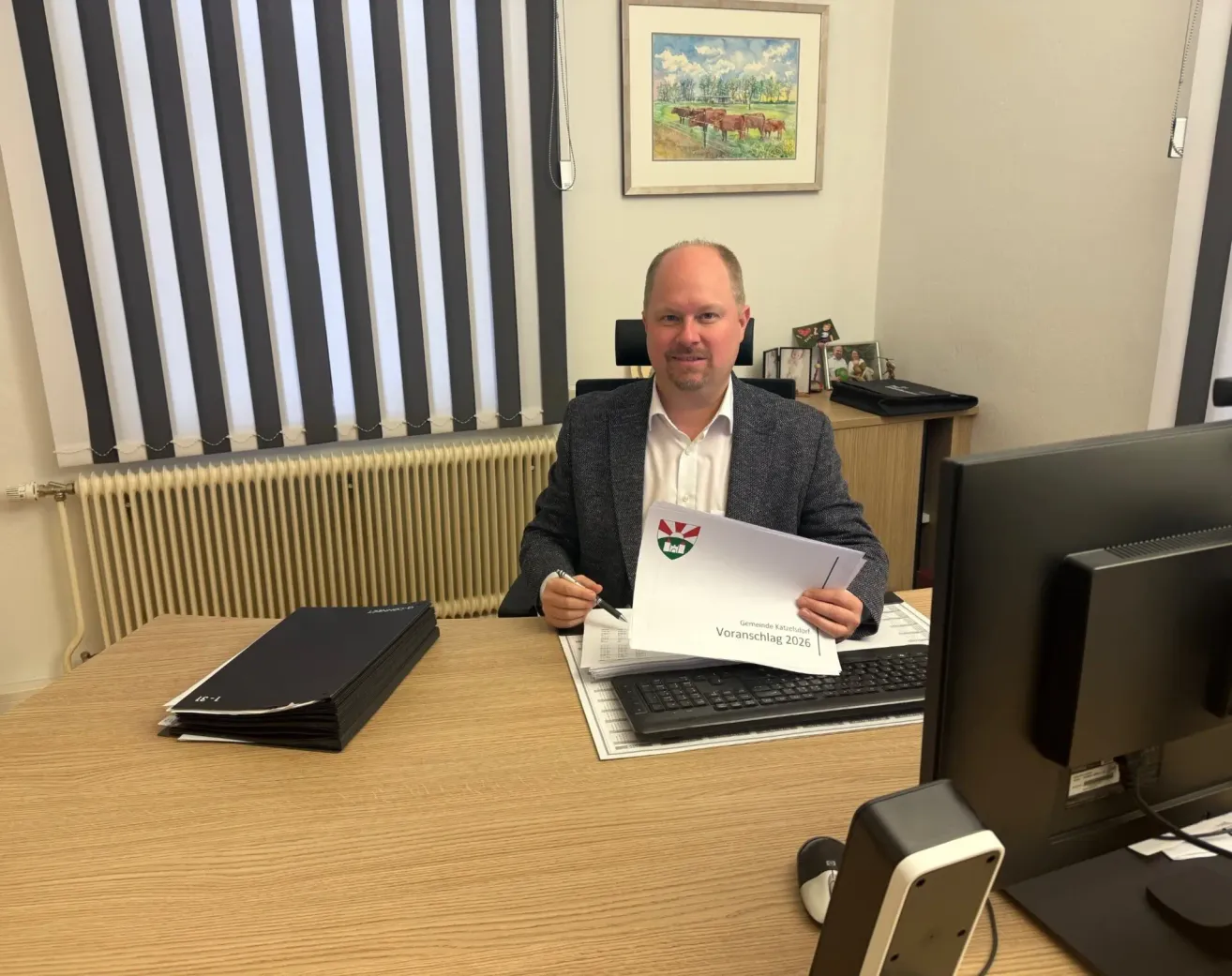 Ein Mann in Anzug sitzt an einem Schreibtisch mit einer deutschen Zeitung mit dem Titel 'Voranschlag 2026'. Hinter ihm ist ein Schreibtisch mit Computer, Tastatur und Monitor. Links ist ein Stapel von Ordnern. Ein Heizkörper ist an der Wand und ein Bilderrahmen ist darüber montiert.