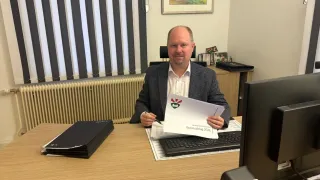 Ein Mann in Anzug sitzt an einem Schreibtisch mit einer deutschen Zeitung mit dem Titel 'Voranschlag 2026'. Hinter ihm ist ein Schreibtisch mit Computer, Tastatur und Monitor. Links ist ein Stapel von Ordnern. Ein Heizkörper ist an der Wand und ein Bilderrahmen ist darüber montiert.