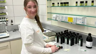 Eine Frau in einem weißen Laborkittel steht in einem Labor, lächelt, mit Regalen verschiedener Flaschen hinter ihr und einem Tresen mit mehreren Flaschen und einer Waage vor ihr.