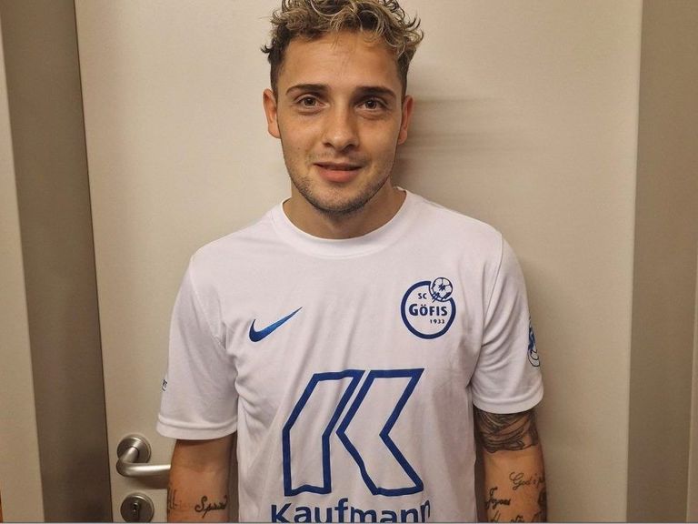 Ein Mann mit blondem Haar und Bart trägt ein weißes Nike-Shirt mit blauem Logo auf der linken Seite und dem Wort Kaufmann auf der rechten Seite.