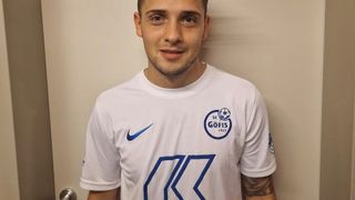 Ein Mann mit blondem Haar und Bart trägt ein weißes Nike-Shirt mit blauem Logo auf der linken Seite und dem Wort Kaufmann auf der rechten Seite.