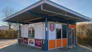 Ein Imbiss mit einer Speisekarte steht unter einem Metalldach. Der Kiosk ist orange und weiß gestrichen, und draußen stehen Tische und Stühle.