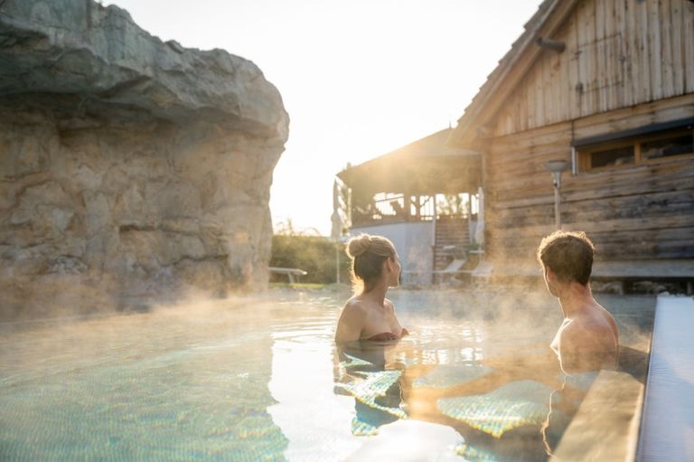 Ein Mann und eine Frau sitzen in einem Pool und genießen die Aussicht auf ein Holzhaus und eine große Felsformation unter einem strahlenden Himmel.
