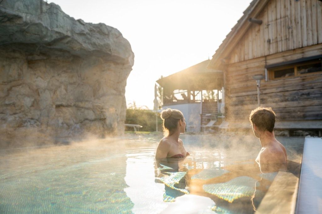 Ein Mann und eine Frau sitzen in einem Pool und genießen die Aussicht auf ein Holzhaus und eine große Felsformation unter einem strahlenden Himmel.