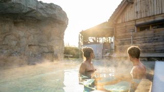 Ein Mann und eine Frau sitzen in einem Pool und genießen die Aussicht auf ein Holzhaus und eine große Felsformation unter einem strahlenden Himmel.