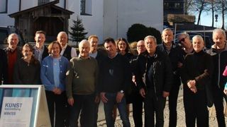 Eine Gruppe von Menschen posiert für ein Foto vor einer Kirche, einige mit Brille und lächelnd, mit einem Schild, das Kunstausstellung Winter steht.
