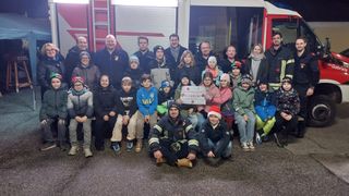Eine Gruppe von Menschen, einschließlich Feuerwehrleute, Erwachsene und Kinder, posiert für ein Foto vor einem Feuerwehrwagen. Ein Banner mit dem Text € 2.603,50 ist sichtbar.