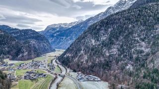 Ein bergiges Tal mit schneebedeckten Gipfeln, einem gewundenen Fluss und einem kleinen Dorf mit Häusern und einer Straße.