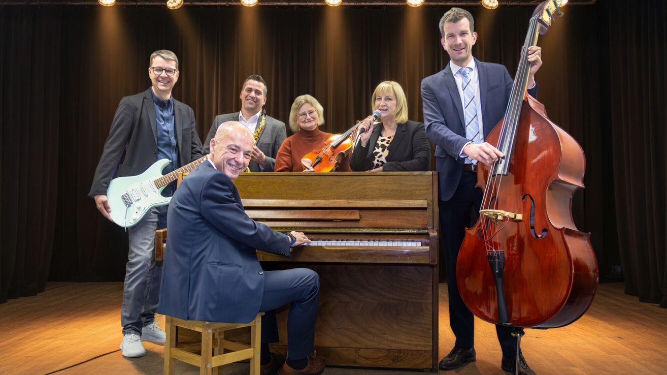 Eine Gruppe von Musikern posiert für ein Foto in einem Studio. Ein Mann spielt Klavier, eine Frau hält Geige und Mikrofon. Ein anderer Mann hält ein Saxophon.