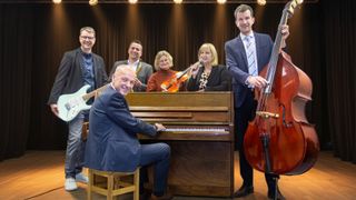 Eine Gruppe von Musikern posiert für ein Foto in einem Studio. Ein Mann spielt Klavier, eine Frau hält Geige und Mikrofon. Ein anderer Mann hält ein Saxophon.