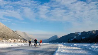 Vier Personen eislaufen auf einem gefrorenen See, tragen Helme und Winterkleidung. Sie fahren in einer Reihe, mit Bergen und blauem Himmel im Hintergrund.