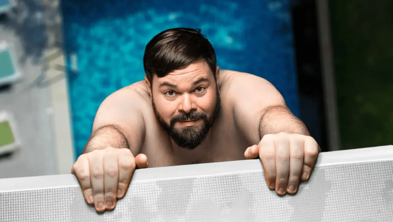 Ein Mann mit Bart lehnt sich an den Rand eines Schwimmbeckens, seine Augen auf etwas vor ihm gerichtet. Er ist oberseits ohne Kleidung, sichtbare Muskeln. Das Becken ist blau.