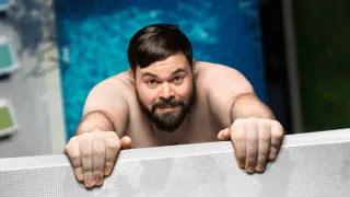 Ein Mann mit Bart lehnt sich an den Rand eines Schwimmbeckens, seine Augen auf etwas vor ihm gerichtet. Er ist oberseits ohne Kleidung, sichtbare Muskeln. Das Becken ist blau.