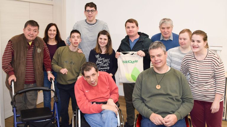 Eine Gruppe von Menschen posiert für ein Foto in einem Raum. Ein Mann im Rollstuhl hält eine WaldQuelle-Tasche.