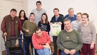 Eine Gruppe von Menschen posiert für ein Foto in einem Raum. Ein Mann im Rollstuhl hält eine WaldQuelle-Tasche.