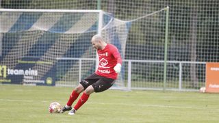 Ein Fußballspieler in einem roten Trikot tritt einen Ball auf einem Feld, hinter ihm steht ein Tornetz.