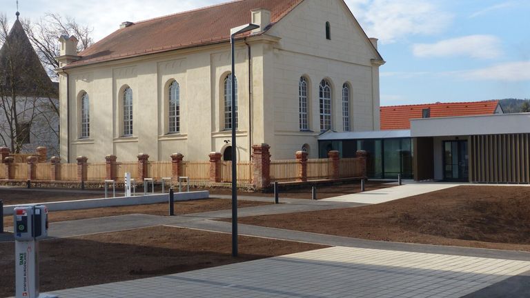Eine alte Kirche mit rotem Dach und beigefarbenen Mauern. Sie hat bogenförmige Fenster und einen Zaun davor.