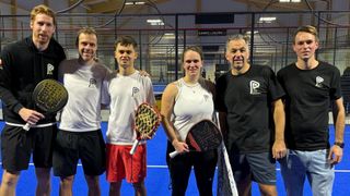 Vier Personen stehen auf einem Paddle-Tennisplatz und lächeln für ein Foto. Zwei von ihnen halten Schläger. Der Platz hat einen blauen Boden und einen schwarzen Zaun. Der Mann rechts trägt ein schwarzes T-Shirt. Der Mann links trägt ein weißes T-Shirt.