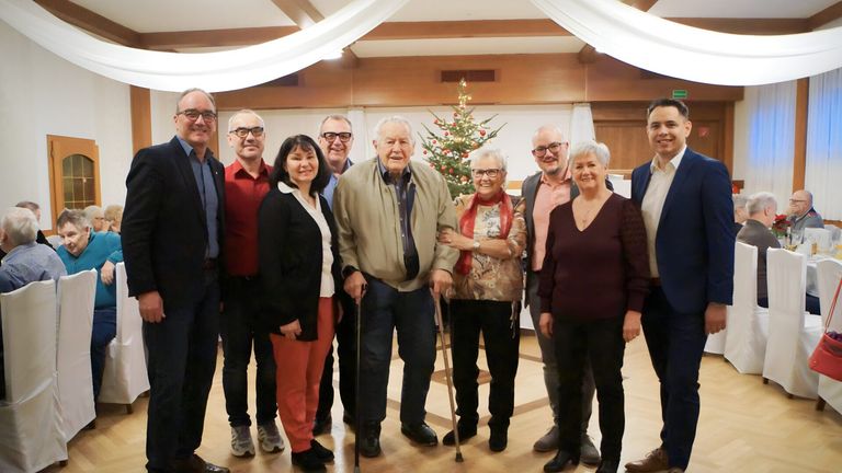 Eine Gruppe von Menschen, darunter Männer und Frauen, steht gemeinsam in einem Raum und lächelt für ein Foto. Ein Weihnachtsbaum steht im Hintergrund.