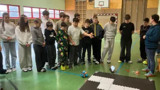 In einer überdachten Basketballhalle versammelt sich eine Gruppe von Jungen um einen kleinen Roboter auf dem Boden, wahrscheinlich beschäftigt mit einer Robotik-Aktivität.