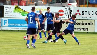 Fünf Fußballspieler spielen auf einem Feld. Zwei Spieler in blauen Trikots, einer mit der Nummer 6, stehen einem Spieler in Schwarz gegenüber. Dahinter beobachten zwei andere. Im Hintergrund befindet sich eine Wand mit Werbung.