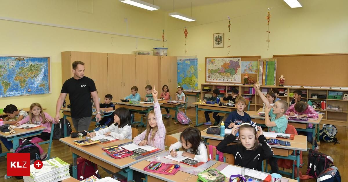 Ein Klassenzimmer mit Schülern an Schreibtischen, ein Lehrer steht vorne. Der Raum hat helle Holzschränke, eine Karte an der Wand und verschiedene Lehrmaterialien auf den Schreibtischen.
