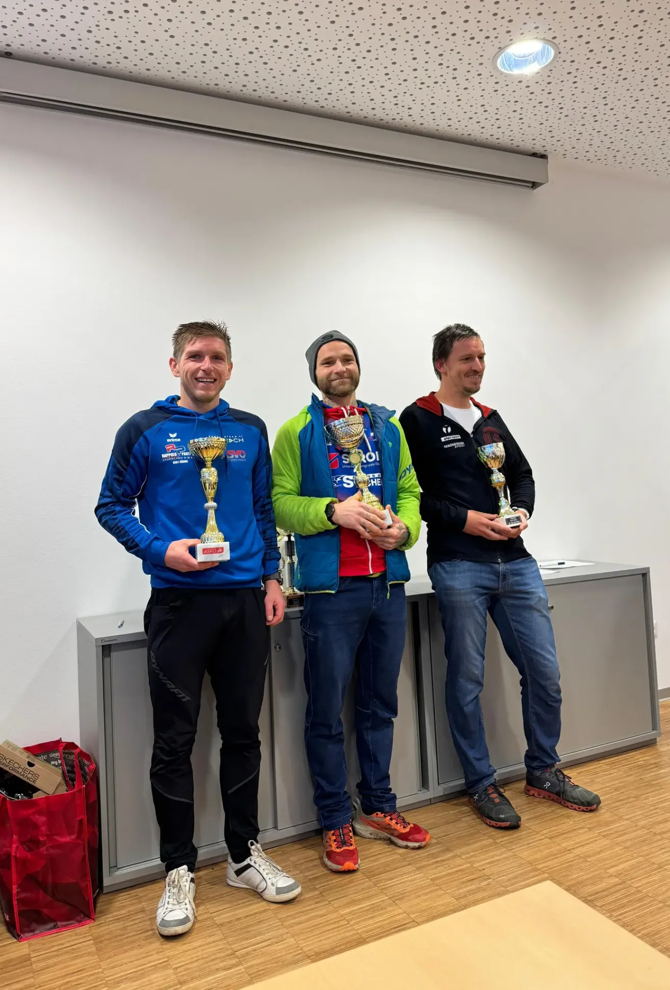 Drei Männer stehen zusammen, halten Trophäen in der Hand und stehen vor einer weißen Wand. Sie lächeln und sind in Sportbekleidung gekleidet. Der Mann links trägt eine blaue Jacke, der in der Mitte eine grüne Jacke und der rechts eine schwarze Jacke. Hinter ihnen steht ein grauer Schrank.