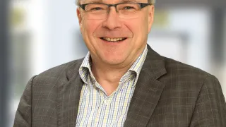 Ein lächelnder Mann in einem grauen Anzug und mit Brille steht in einem Raum mit verschwommenem Hintergrund, vielleicht für ein professionelles Foto.