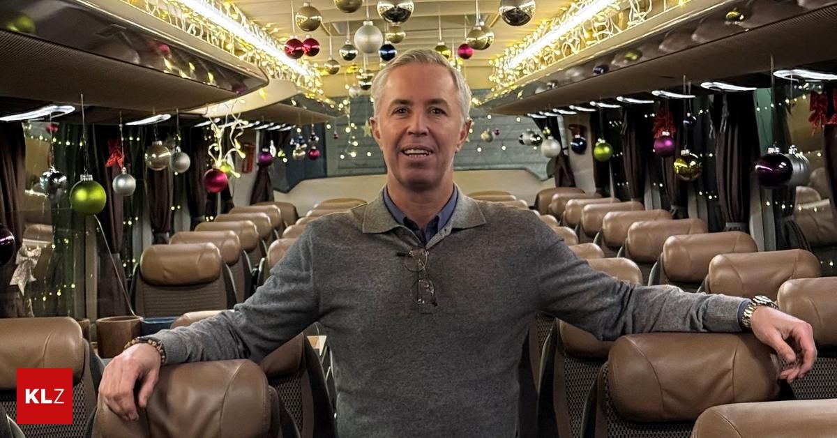 Ein Mann steht lächelnd in einem dekorierten Bus und trägt einen grauen Pullover. Der Bus hat Ledersitze und Weihnachtskugeln hängen von der Decke.