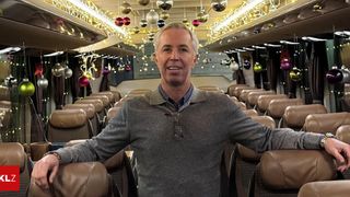 Ein Mann steht lächelnd in einem dekorierten Bus und trägt einen grauen Pullover. Der Bus hat Ledersitze und Weihnachtskugeln hängen von der Decke.
