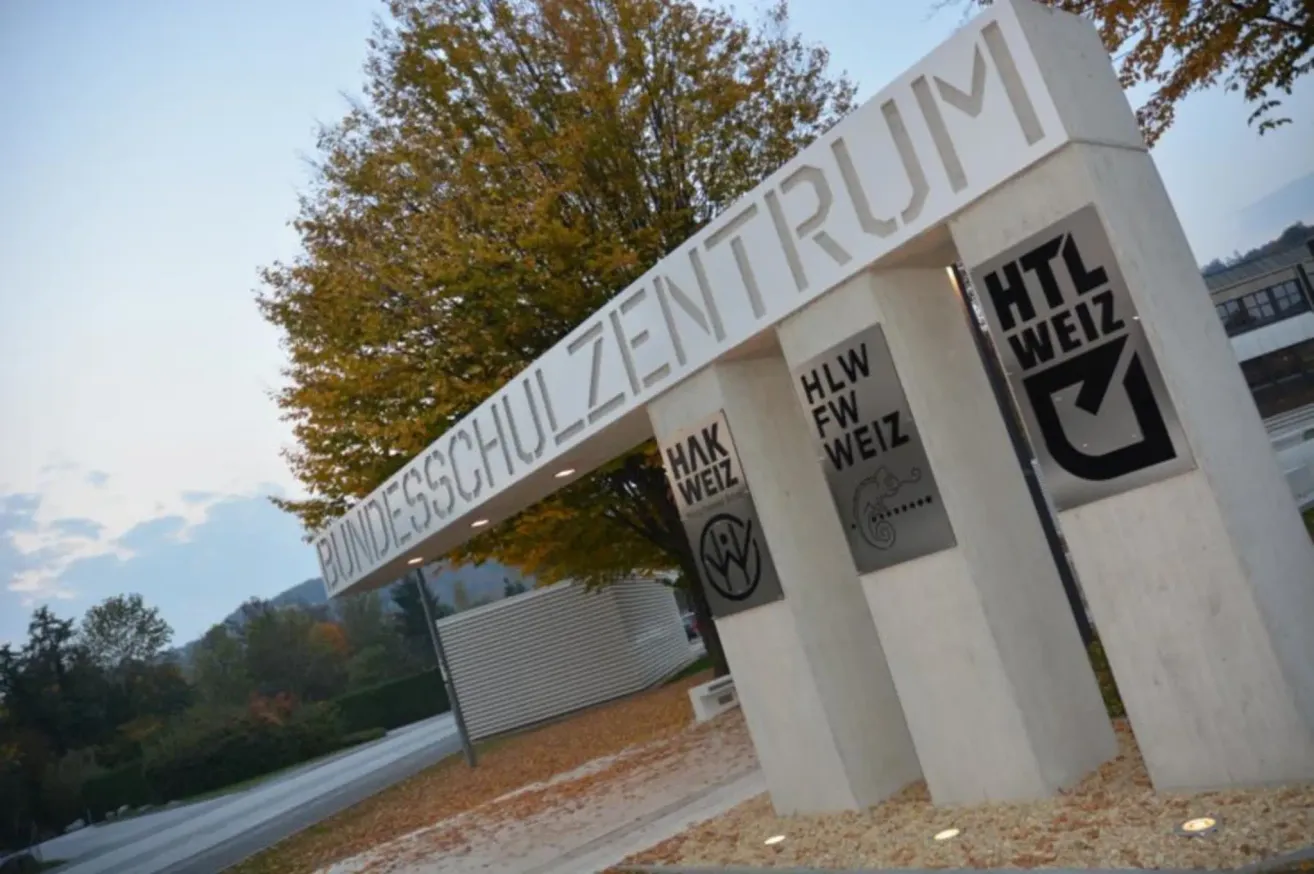 Ein Eingangsschild für Bundesschulzentrum mit HLW FW Weiz und HAK Weiz an den Seiten, unter einem Baum mit Herbstlaub.
