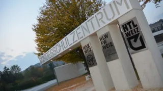 Ein Eingangsschild für Bundesschulzentrum mit HLW FW Weiz und HAK Weiz an den Seiten, unter einem Baum mit Herbstlaub.