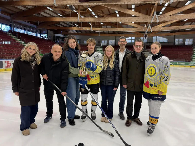 Eine Gruppe von Hockeyspielern in Uniformen, einer hält einen Pokal, steht auf einer Eisbahn mit Erwachsenen, darunter ein Mann mit Brille, und leeren Tribünen im Hintergrund.