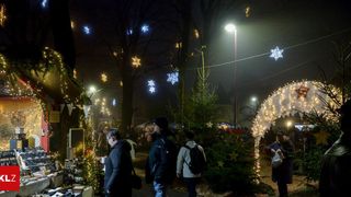 Menschen gehen auf einem festlich geschmückten Außenmarkt, mit Bäumen, die mit Ornamenten und Lichtern geschmückt sind. Ein Mann mit Rucksack und eine Frau mit Plastiktüte gehören zur Menge.