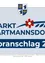 Das Bild zeigt ein Poster für 'Markt Hartmannsdorf' mit einem Entwurf für 2023. Der Wappenschild von Weiz ist oben abgebildet, gefolgt von einer Blume und dem Text. Darunter wird der Bevölkerungsstand vom 31. Oktober 2024 mit 2.992 Einwohnern angegeben.