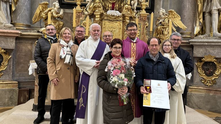 Eine Gruppe von Menschen, einschließlich Geistlicher, steht in einer Kirche. Eine Frau hält Blumen und ein Zertifikat, während ein Priester lächelt. Hinter ihnen sind Statuen und Kerzen zu sehen.