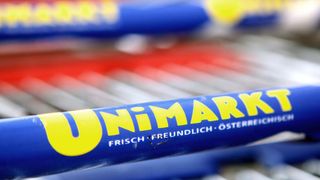 Ein Nahaufnahme eines blauen Einkaufswagengriffs, der mit UniMARK beschriftet ist, mit verschwommenen bunten Artikeln im Hintergrund.