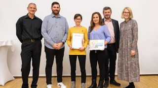 Eine Gruppe von sechs Personen steht gemeinsam in einem Raum, wahrscheinlich für eine Preisverleihung. Die Frau in der Mitte hält ein Zertifikat und ein Buch, während andere lächeln.