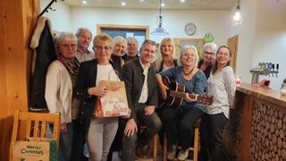 Eine Gruppe Erwachsener versammelt sich in einem Raum und lächelt für ein Foto. Eine Frau hält einen Geschenkbeutel mit Weihnachtsthema. Eine andere Frau spielt eine Gitarre. Der Raum hat einen Holzrahmen um ein Fenster und eine froschförmige Uhr.