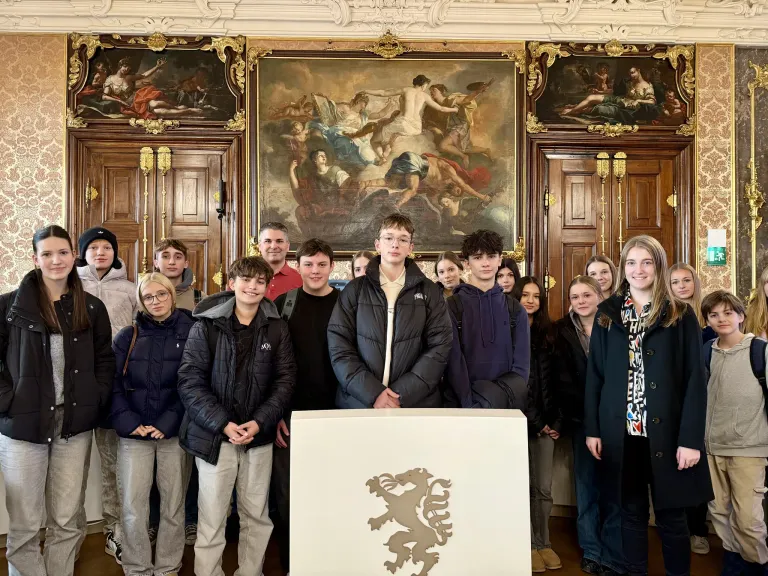 Eine Gruppe von Schülern und Erwachsenen steht in einem eleganten Raum und lächelt für ein Foto. Ein großes Gemälde ist an der Wand hinter ihnen.