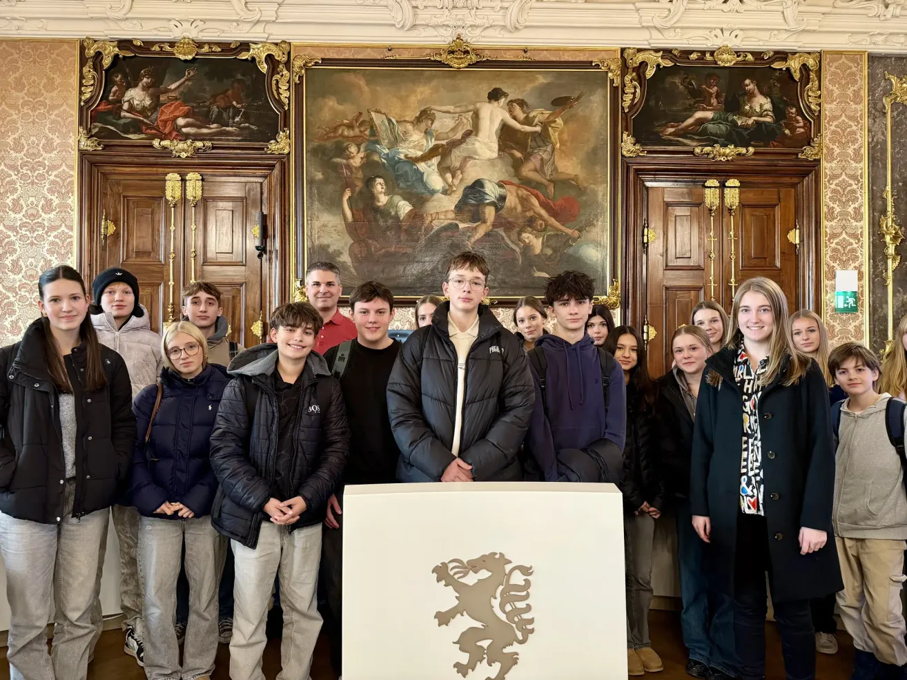 Eine Gruppe von Schülern und Erwachsenen steht in einem eleganten Raum und lächelt für ein Foto. Ein großes Gemälde ist an der Wand hinter ihnen.